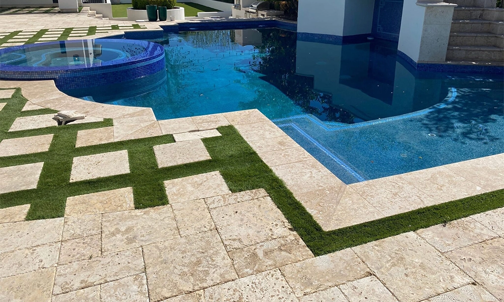 A travertine pool deck with a unique pattern of grass inlays, surrounding a pool with a blue tiled hot tub and steps in the background, creating a modern and natural outdoor aesthetic