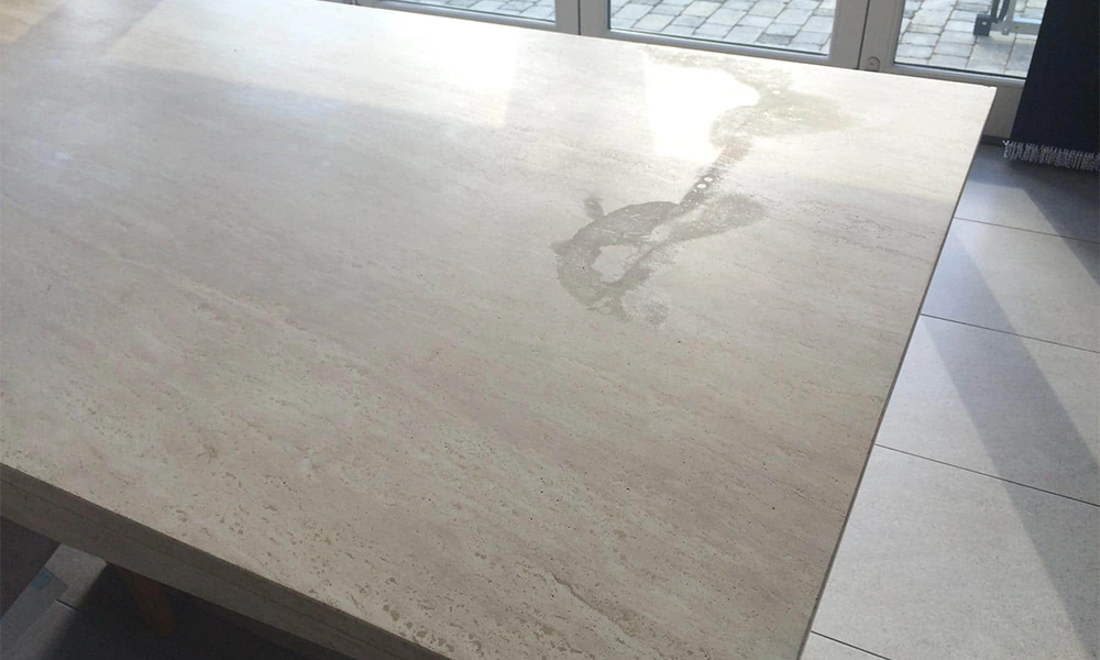 "Travertine tabletop with visible water stain marks under sunlight near glass doors."