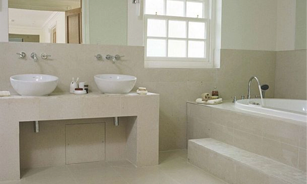Minimalistic bathroom featuring beige stone tiles, a freestanding bathtub, and a sleek double sink vanity with wall-mounted faucets.