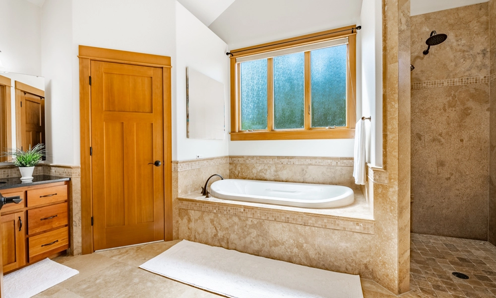 A spacious bathroom featuring travertine stone walls and floor, a separate shower, a bathtub area with a window providing natural light, and wooden door frames adding warmth to the space.